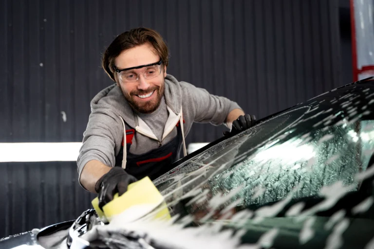 A smiling male mechanic in glasses and a uniform polishing a car windshield in a workshop.